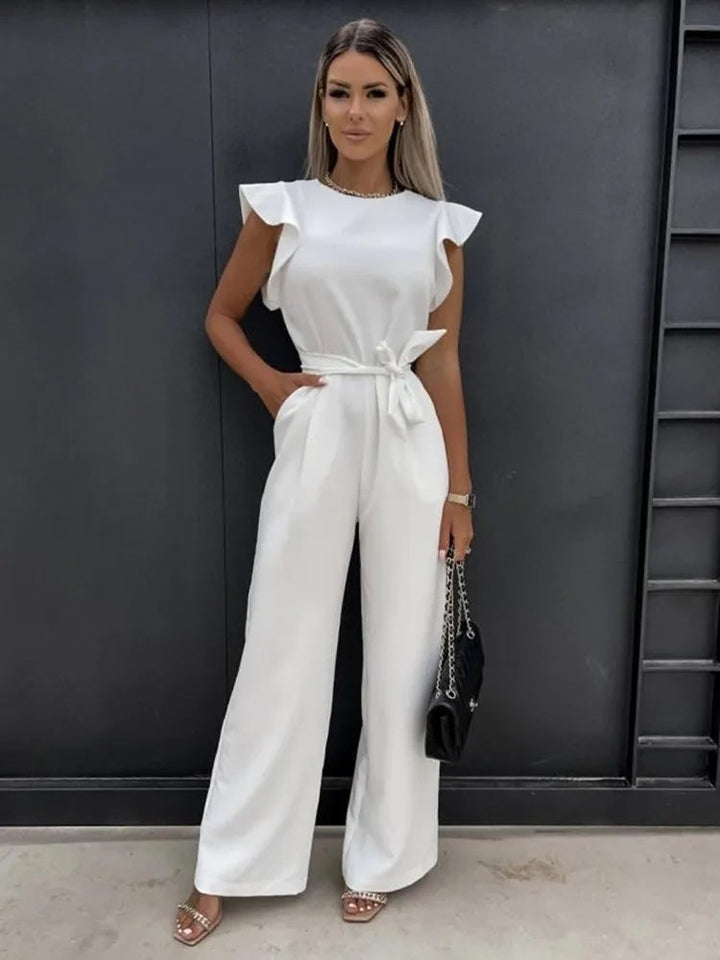 Femme debout portant une combinaison blanche moderne à coupe large, sans manches, avec épaules structurées, taille nouée et pantalon fluide, offrant une allure élégante et raffinée, accessoirisée d’un sac à main noir et de sandales à talons.